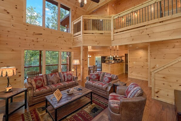A spacious log cabin interior with a high ceiling, a central seating area with two sofas and a coffee table, a kitchen area, and a balcony with a railing.