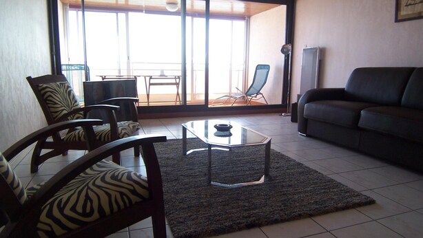 A living room with a sofa, chairs, a coffee table, and a view through a glass door to an outdoor area with a chair and a table. - Apartment with canal view, garage and wifi (Les Sables-d'Olonne)