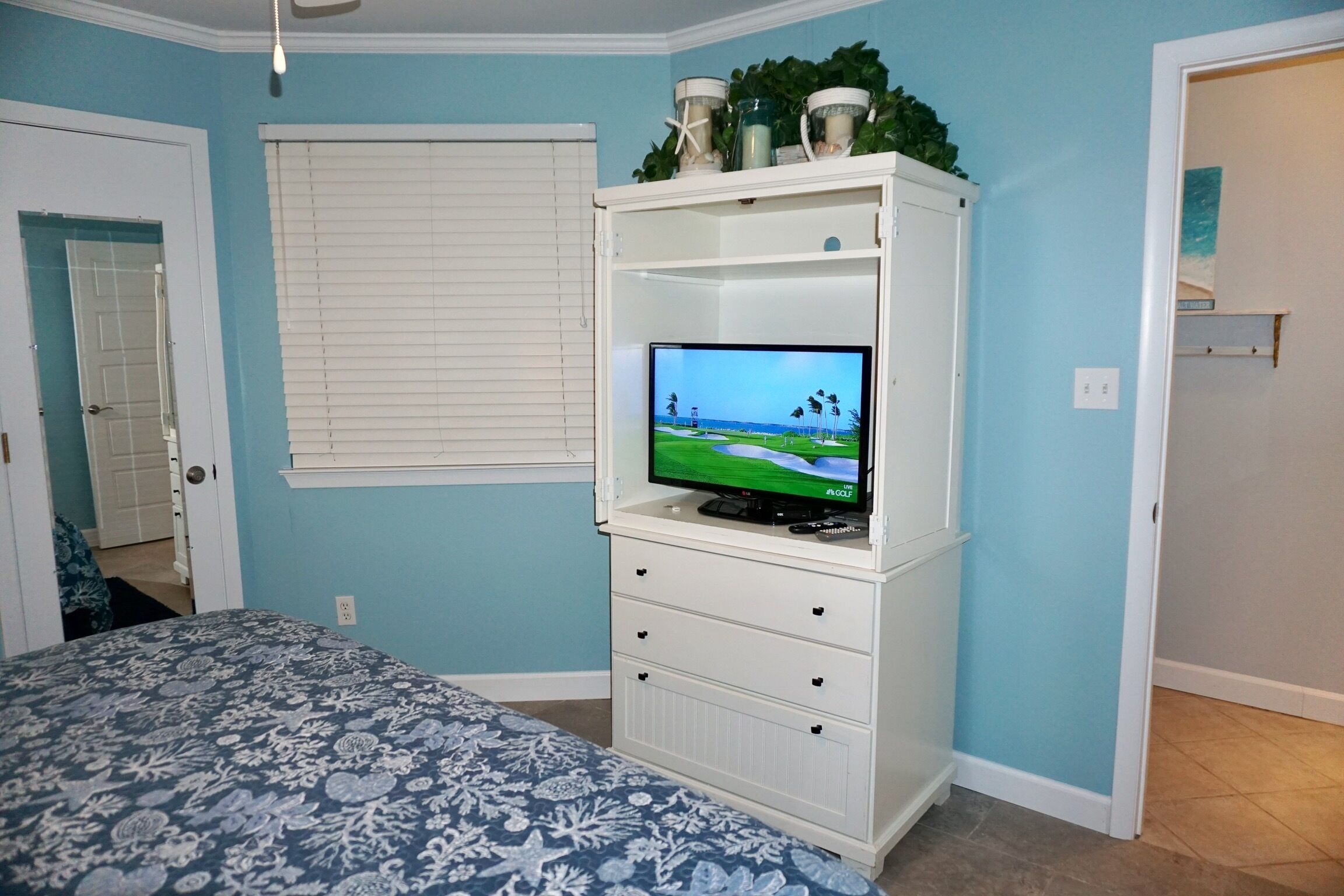 Een slaapkamer met een bed, een televisie op een wit kastje, een raam met zonwering, en een deur.