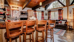 A rustic cabin interior with wooden furniture, a kitchen area, and large windows offering views of the outdoors.