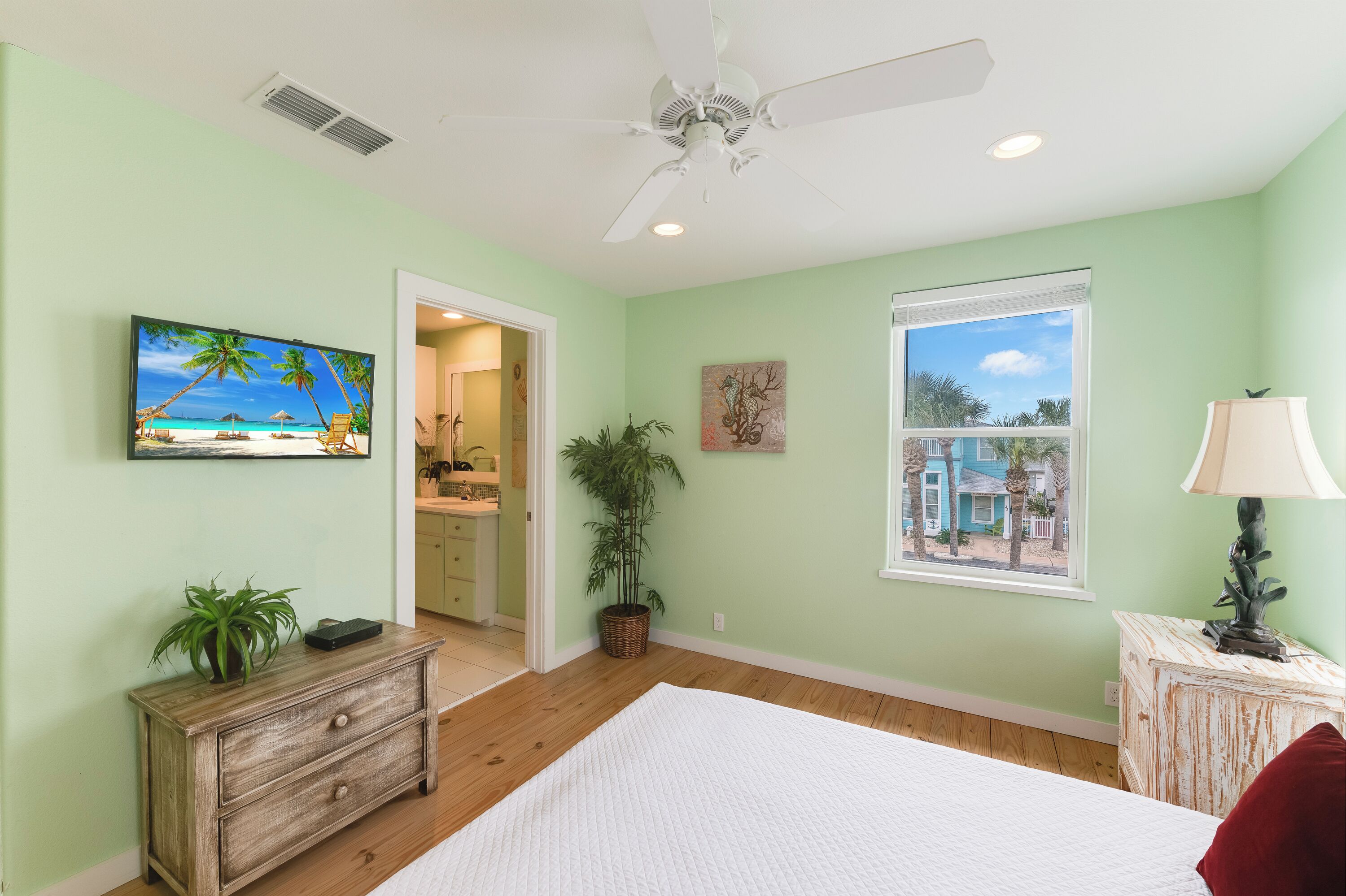 Une chambre à coucher avec un lit, un ventilateur de plafond, une fenêtre donnant sur un beau paysage, une commode, une plante et un tableau.