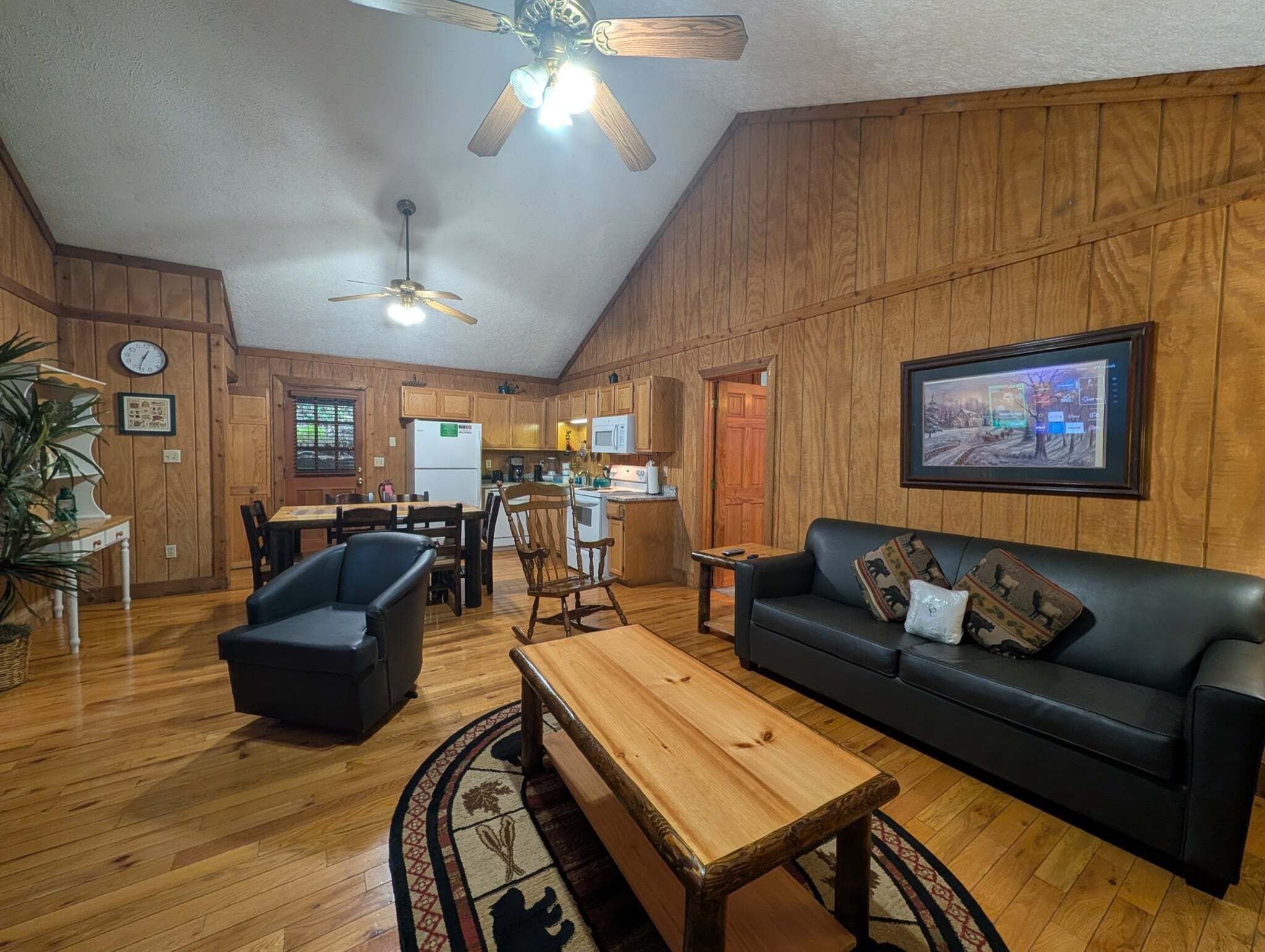 A cozy living room with a wooden floor, a black leather sofa, a wooden coffee table, a dining table with chairs, a kitchen area, and a ceiling fan.