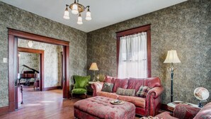 A spacious living room with a red sofa, green armchair, and a large ottoman. There is a piano in the background, and the room has patterned wallpaper and wooden flooring. - Gorgeous 20s Historic Mini Mansion. King & Queens. Downtown Strip. (Wylie)