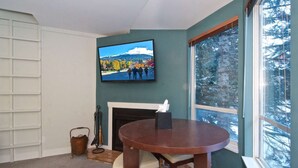A cozy living room with a fireplace, a round table, and a large window with a view of trees. - Cute and Cozy Creekside townhouse. Ground floor, easy access, free parking, 100 meters to lifts (Whistler)