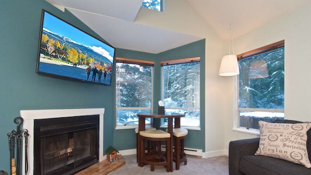 A living room with a fireplace, a TV mounted on the wall, and a view of snowy trees through the windows. - Cute and Cozy Creekside townhouse. Ground floor, easy access, free parking, 100 meters to lifts (Whistler)