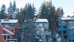 A multi-story building with a red brick facade, a grey upper level, and a blue lower level. There is a staircase connecting the two levels, and a small shed attached to the building. - Cute and Cozy Creekside townhouse. Ground floor, easy access, free parking, 100 meters to lifts (Whistler)