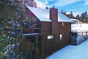 Una cabaña de madera de dos pisos con balcón, rodeada de terreno y árboles cubiertos de nieve.