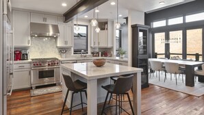 A modern kitchen with white cabinetry, a central island with stools, and a dining area with a table and chairs. - Ski-In/Ski-Out Home, Private Hot Tub (Park City)