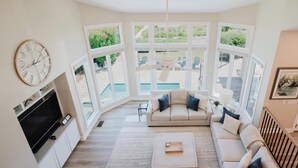 Una sala de estar con sofá, mesa de centro, televisor y ventanas amplias con vista a una piscina y jardín.