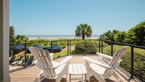 Una terraza con dos sillas Adirondack blancas que dan a una playa, una palmera y un cielo azul despejado.
