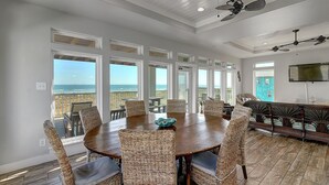 A spacious dining area with a large round table and wicker chairs, overlooking a balcony with a view of the ocean.