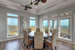 A dining area with a round table and wicker chairs, large windows with ocean views, and a ceiling fan.