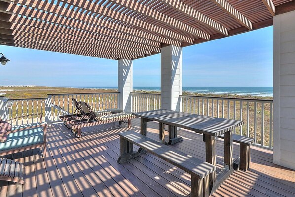 Eine Holzterrasse mit Pergola, Tisch und Bank mit Blick auf einen Strand.