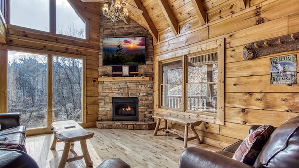 A cozy cabin interior with a stone fireplace, wooden walls, and large windows offering a view of the forest.