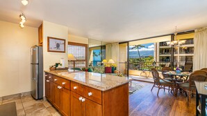 A kitchen with wooden cabinets and a large island, a dining area with wicker chairs, and a balcony with a view of the ocean.