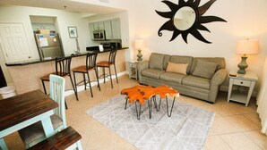 A living room with a sofa, two chairs, a coffee table, and a rug. There is a kitchen area in the background with cabinets and appliances.