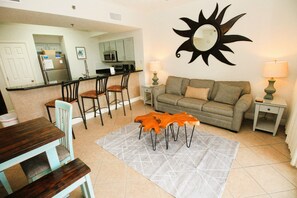 Sala de estar con un sofá, dos sillones, una mesa de centro y una alfombra. Se observa una cocina al fondo con armarios y electrodomésticos.