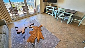 A spacious living area with a large window overlooking a beach, a flat-screen TV mounted on the wall, and a wooden coffee table.