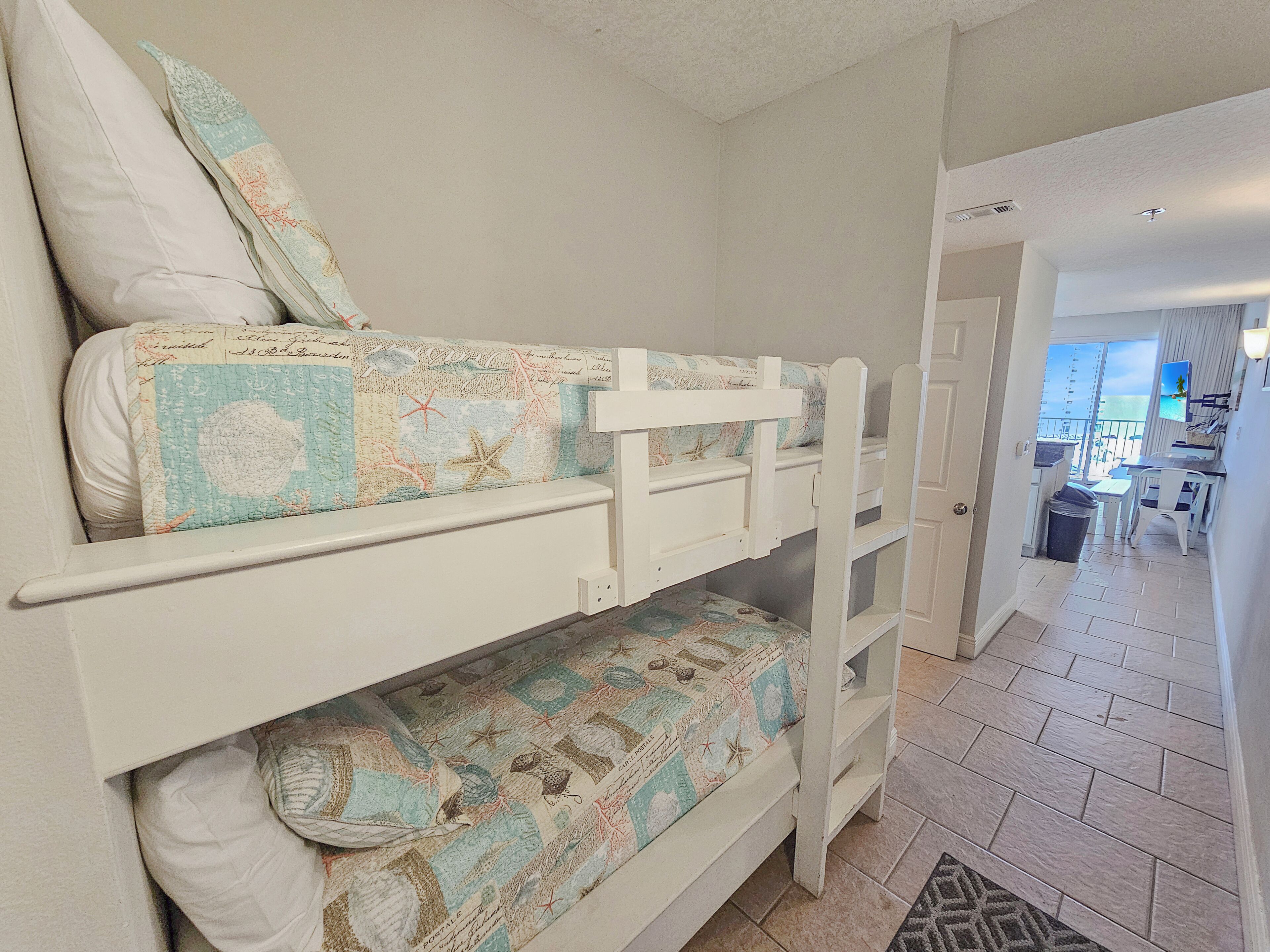 Una cama individual con ropa de cama temática náutica, cabecera blanca y una pequeña zona de escritorio con una silla y un cubo de basura.