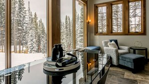 A cozy room with large windows offering a snowy forest view, a glass table with binoculars and a book, and a patterned armchair. - Jane's Ski Chalet | Luxury Mountain Mansion | Sleeps 17 | Modern Decor | Private Hot Tub (Winter Park)