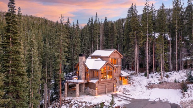 A log cabin with a stone chimney, large windows, and a deck, surrounded by a dense forest and snow. - Jane's Ski Chalet | Luxury Mountain Mansion | Sleeps 17 | Modern Decor | Private Hot Tub (Winter Park)