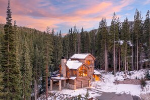Una cabaña de troncos con chimenea de piedra, ventanas amplias y una terraza, rodeada por un bosque denso y nieve.