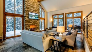 A spacious living room with a stone fireplace, large windows offering a view of snow-covered trees, and a comfortable seating area. - Jane's Ski Chalet | Luxury Mountain Mansion | Sleeps 17 | Modern Decor | Private Hot Tub (Winter Park)