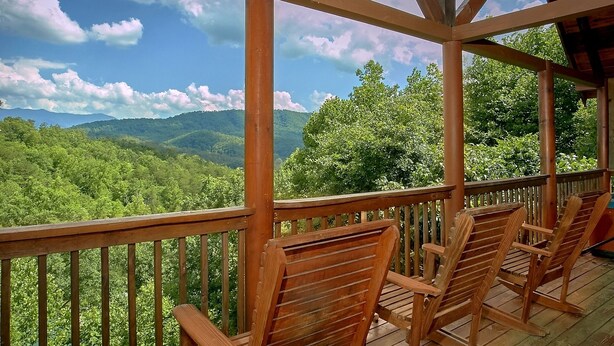 Una terraza de madera con cuatro sillas de madera y una barandilla, con vistas a un valle boscoso y montañas al fondo.