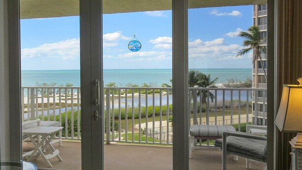 A balcony with a view of the ocean, white outdoor furniture, and a palm tree. - Escape To Beachfront Getaway Vacation Condo At Bargain Price! Heated Resort Pool! (Fort Myers Beach)