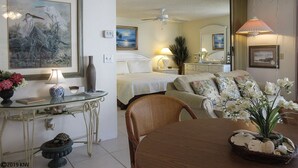 A well-lit room with a bed, a dining table with chairs, a small table with a lamp, and a wall-mounted TV. - Escape To Beachfront Getaway Vacation Condo At Bargain Price! Heated Resort Pool! (Fort Myers Beach)