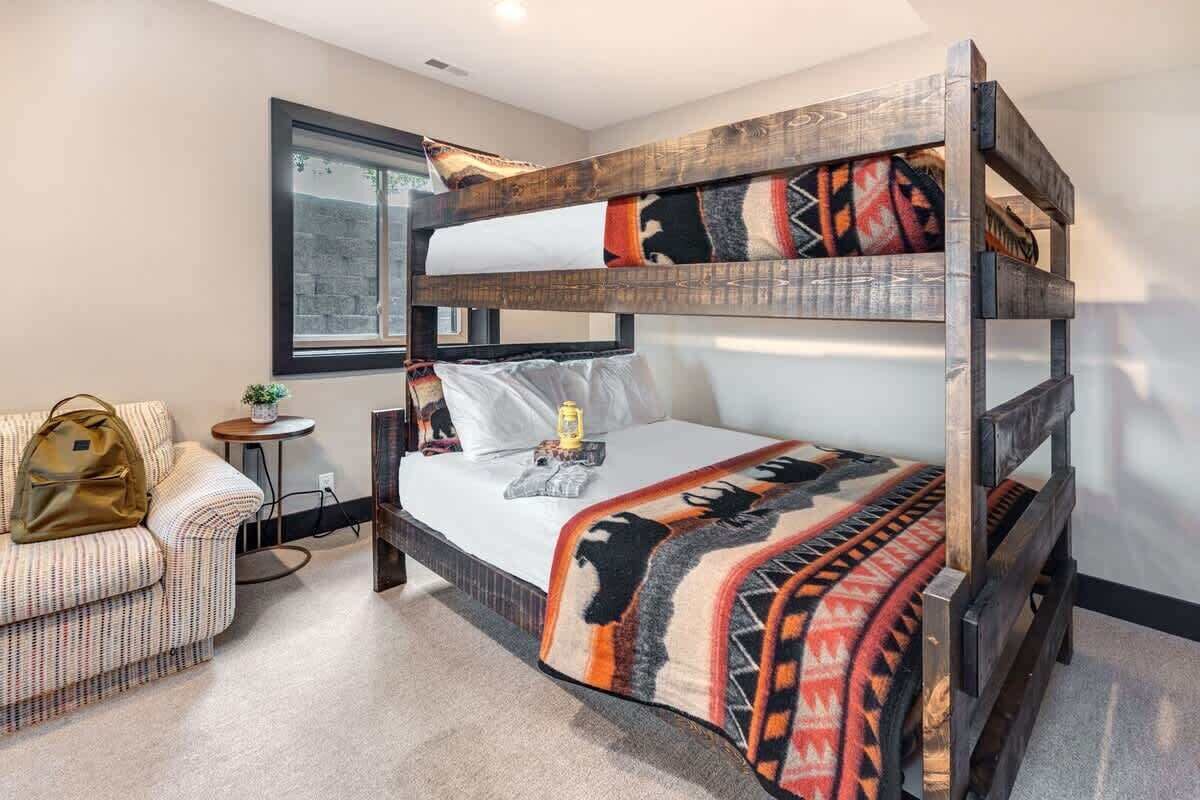 A bunk bed with a patterned blanket, a small round table with a plant, and a couch with a bag.
