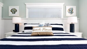 Un lit bien fait, avec une literie à rayures et un coussin sur lequel est écrit « Maison de plage ».