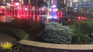 A nighttime view of a fountain with colorful lights, a stone wall, and a building with illuminated signage in the background.