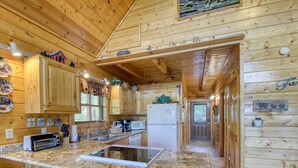 A wooden cabin with a kitchen featuring granite countertops, a built-in microwave, and a refrigerator. There are wooden stools and a window with curtains.
