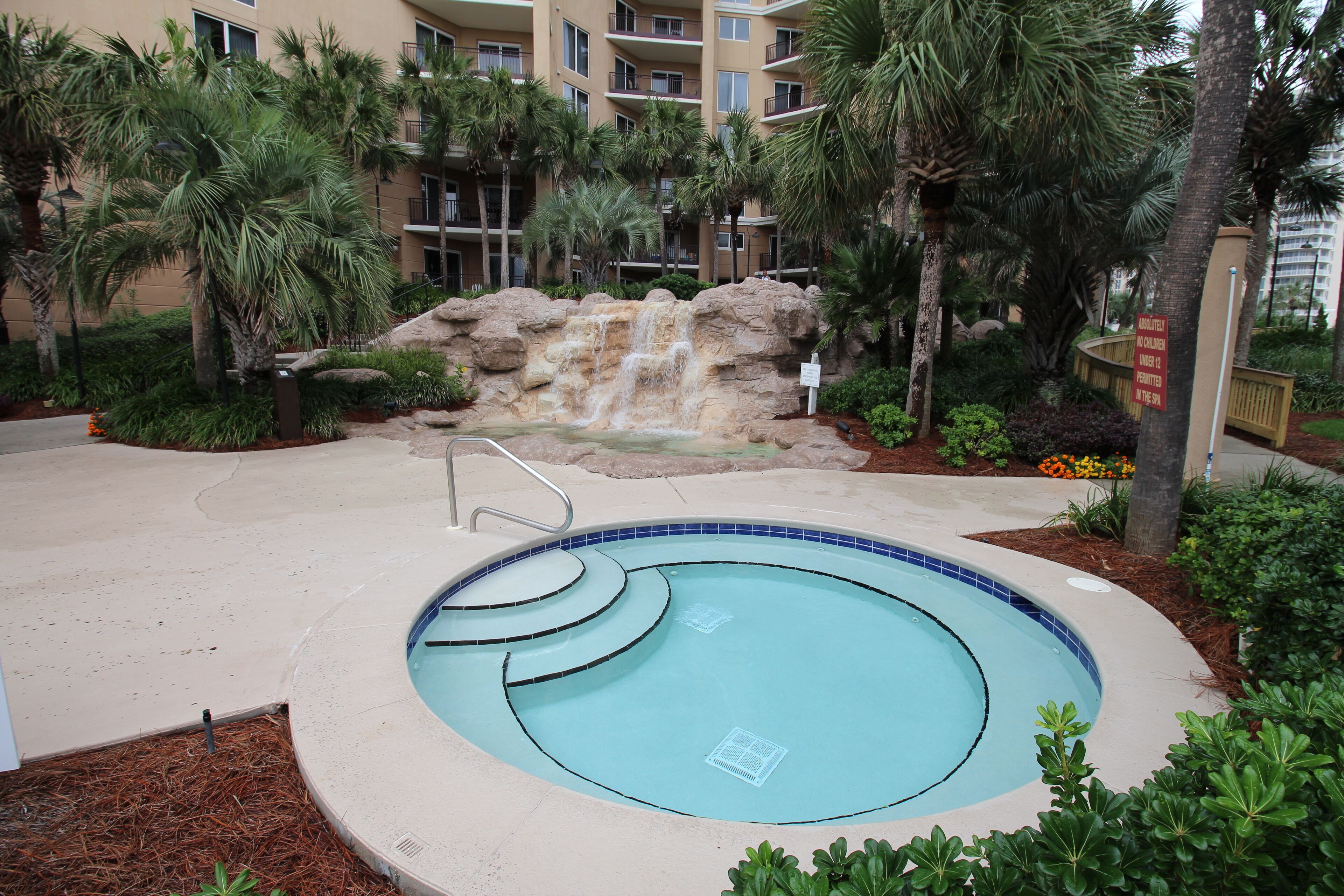 A swimming pool with a waterfall feature, surrounded by palm trees and a multi-story building in the background.
