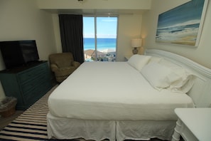 A hotel room with a large bed, a television on a dresser, a chair, a lamp, and a painting of a beach.