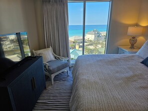 A hotel room with a large window, a bed, a chair, a dresser, and a nightstand with a lamp.