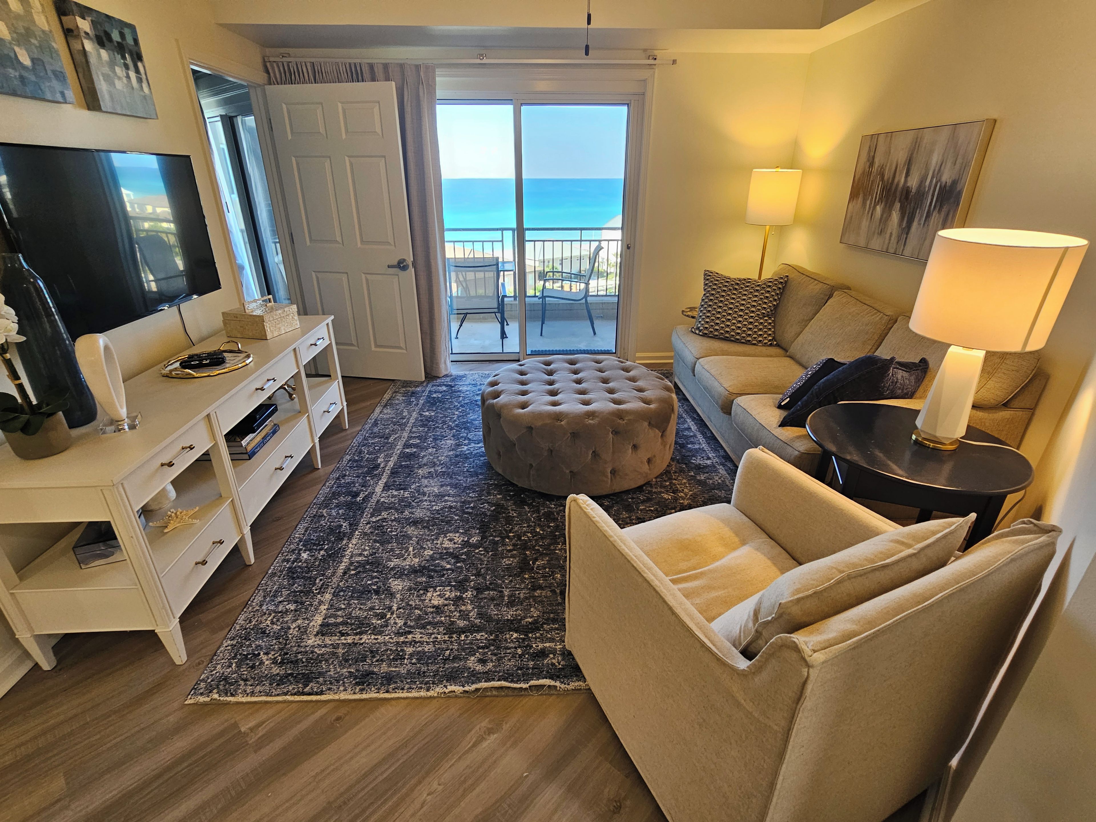 A living room with a television, sofa, ottoman, and a view of the ocean through a sliding door.