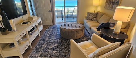 A living room with a television, sofa, ottoman, and a view of the ocean through a sliding door.