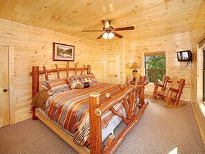 Ein Schlafzimmer in einer Blockhütte mit einem hölzernen Bett, Deckenventilator, Fernseher und Fenster mit Blick ins Freie.