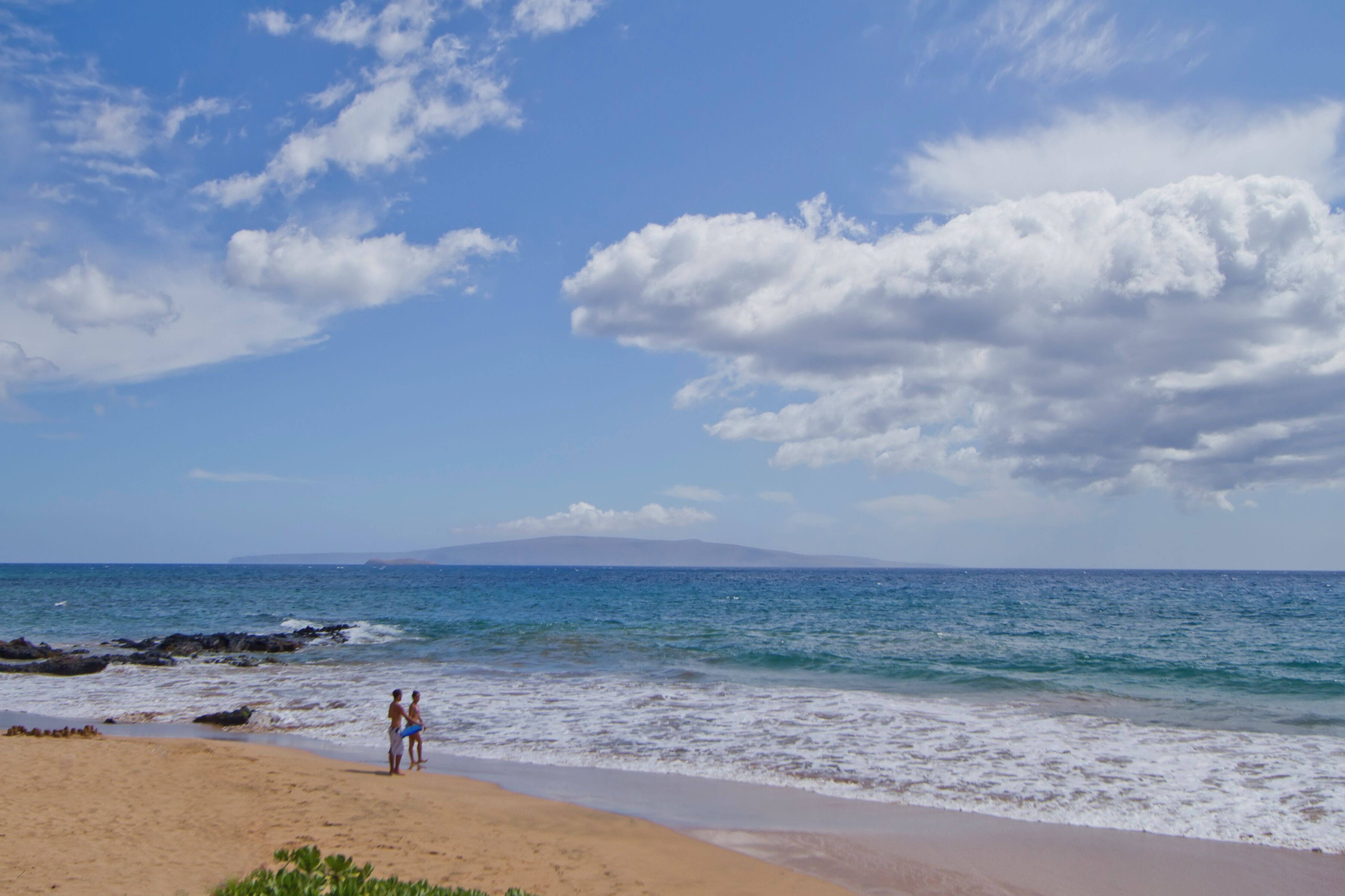 A beach with two people standing near the water's edge, a clear sky, and a distant mountain.