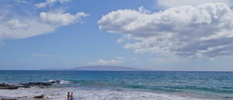 A beach with two people standing near the water's edge, a clear sky, and a distant mountain.