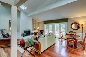 Relax in this spacious living room with a fireplace, sofa set, and dining area, surrounded by lush greenery and a scenic outdoor view.