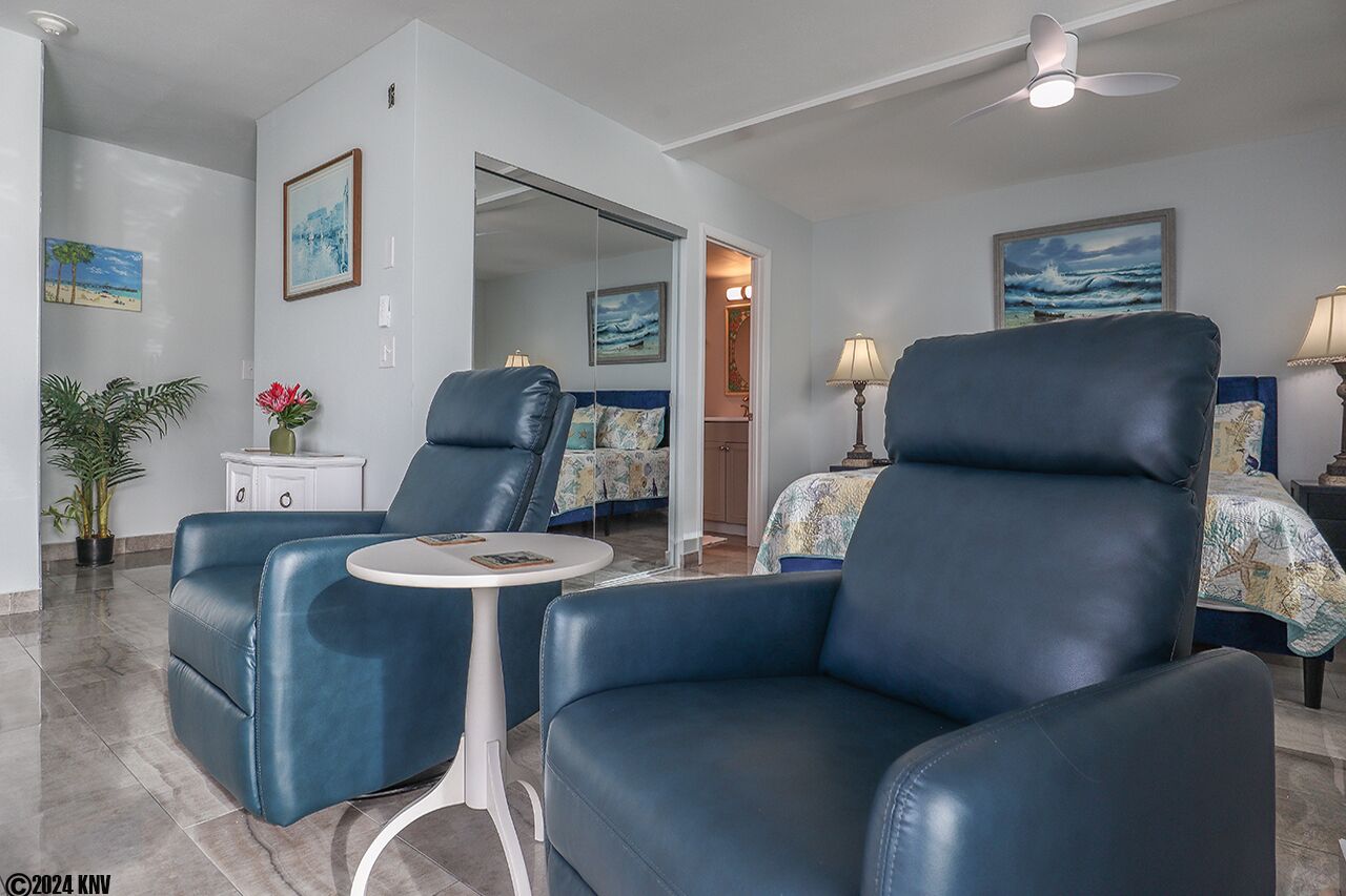 A modern living room with two blue leather armchairs, a small round table, and a bed with a floral bedspread.