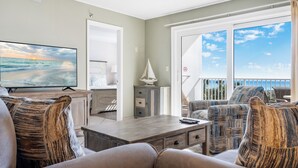 Una sala de estar con televisor, una pequeña decoración de velero y un balcón con vista al océano.