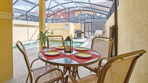 Ein überdachter Essbereich im Freien mit einem Glastisch und Stühlen, ein Pool im Hintergrund und Blick auf benachbarte Gebäude.