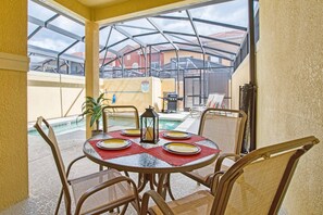 A covered outdoor dining area with a glass table and chairs, a pool in the background, and a view of neighboring buildings.