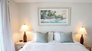 Hotelkamer met een groot bed, twee bedlampen, een schilderij van een tropisch landschap en een raam met gordijnen.