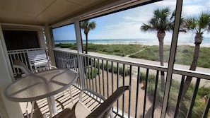 A balcony with a table and chairs, overlooking a beach with palm trees and the ocean. - Island Beach Club 24 - Anna Maria Island - Condo 2 Bedroom / 1 Bath gulf front , maximum occupancy of 4 people. (Holmes Beach)