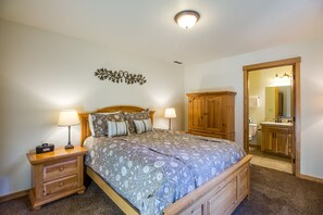 Une chambre à coucher avec un lit, des tables de chevet, une armoire en bois et une salle de bain visible à travers une porte ouverte.
